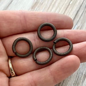 Può includere: Quattro piccoli anellini di giunzione circolari in metallo scuro ossidato. Ogni anello presenta una distinta texture a corda ritorta lungo l'intera circonferenza. Gli anelli sono aperti, con un piccolo spazio nella bobina, e hanno una finitura opaca.