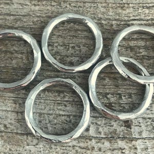 May include: Five silver-colored metal rings with a hammered, textured finish. Each ring features a circular shape with an uneven, organic surface texture. The rings are arranged in a cluster, showcasing the reflective, metallic material and the distinct, hand-crafted appearance of the hammered metal bands.