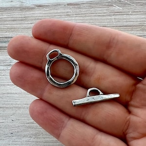 May include: A silver-toned metal toggle clasp set consisting of a circular ring component with a small loop attachment and a separate bar component with a central loop, both featuring a textured, weathered finish and irregular edges.