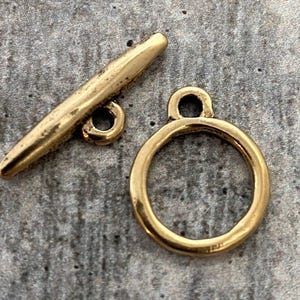 Peut inclure: Un fermoir à bascule doré, composé d'une barre et d'un anneau. La barre est cylindrique avec une extrémité arrondie et une petite boucle. L'anneau est circulaire avec une boucle en haut. Le fermoir est pour bijoux ou accessoires.