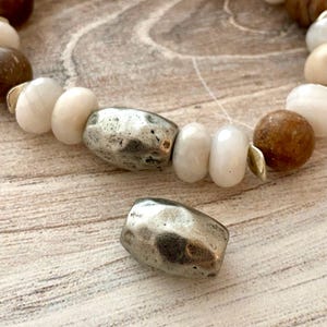 May include: A close-up of a beaded bracelet with a variety of neutral-toned beads. The bracelet features large, hammered silver-colored beads, white oval beads, and brown spherical beads. A single silver bead is separate from the bracelet.
