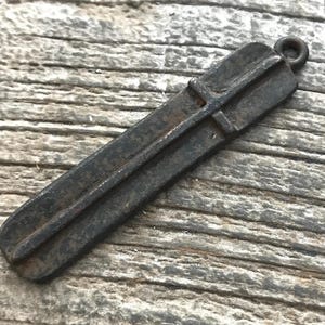 May include: A dark, weathered metal pendant with a long, rectangular shape and a central raised ridge running vertically along its length. The object features a small circular loop at the top end for attachment, displaying a textured, oxidized surface.