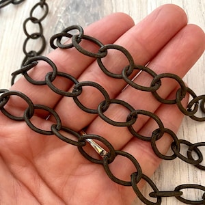 Puede incluir: Primer plano de una cadena de color bronce oscuro con eslabones ovalados. La cadena tiene una superficie texturizada y un cierre dorado. La cadena probablemente se utiliza para joyería o fines decorativos.