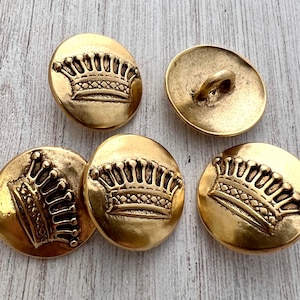 Puede incluir: Cinco botones dorados antiguos con un diseño de corona. Los botones de forma ovalada tienen un lazo en la parte posterior para coser. El diseño de la corona está en relieve en la parte delantera de cada botón.