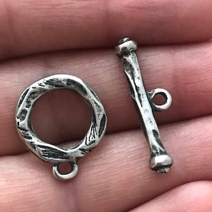 Pode incluir: Close-up de um conjunto de fechos de alternância prateados. O fecho apresenta um anel texturizado e uma barra com um pequeno laço. O metal tem uma aparência envelhecida e rústica, sugerindo uma estética vintage ou artesanal. Ideal para fazer joias.