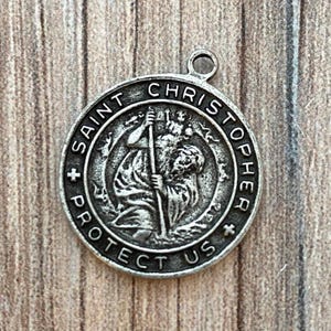 May include: A round silver-toned metal pendant featuring a relief image of Saint Christopher carrying a child across water. The border is inscribed with the text SAINT CHRISTOPHER PROTECT US, accented by two small crosses on the left and right sides.
