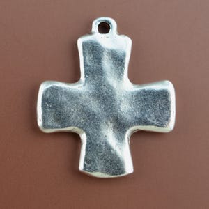 May include: A silver-toned metal cross pendant with a textured, uneven surface and a rounded bail at the top. The cross features wide, flared arms and a smooth, reflective finish that catches the light, creating subtle highlights and shadows.