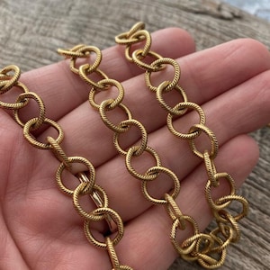 May include: Close-up of a gold-colored chain necklace. The chain links are circular with a textured, rope-like design. The necklace is held in a hand, with a wooden surface in the background.