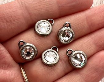 May include: Five small, circular metal charms, each featuring a single clear, faceted crystal set in the center. Each charm has a small loop at the top for attachment. The metal has a dark, slightly weathered finish, contrasting with the bright crystal.