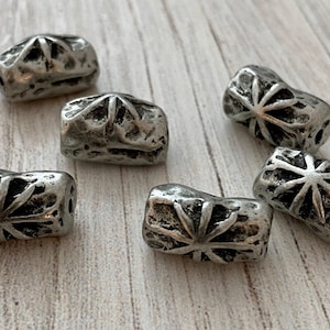 May include: Close-up of six silver-colored, rectangular beads with a starburst design on each face. The beads have a distressed, antique finish and are arranged on a light-colored wooden surface. These beads are suitable for jewelry making.