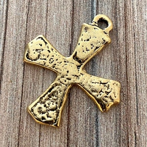 May include: A gold-colored cross pendant with a textured surface. The cross has a loop at the top for hanging and is set against a wooden background. The pendant is a religious symbol.