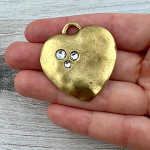 Peut inclure: Pendentif en forme de cœur avec une finition dorée antique texturée. Trois cristaux ronds et transparents sont sertis près du sommet du cœur. Le pendentif a une petite boucle pour l'accrocher. Bijoux.