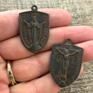 May include: Two dark metal shield-shaped religious pendants. One features a figure with arms outstretched and the text MARIA MEDIADORA ROGAD POR NOS. The other depicts a crucifix with the text S CRISTO DEL AMOR MISERICORDIOSO.