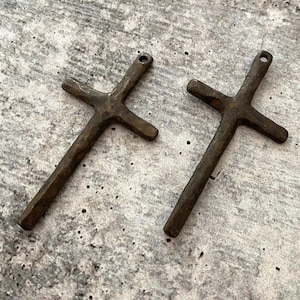 May include: Two rustic, bronze-colored crosses with a textured finish. Each cross has a small hole at the top for hanging. The crosses are displayed on a mottled gray surface.
