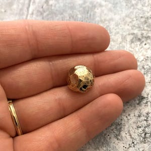 Op de afbeelding: Een kleine, goudkleurige kraal met een facet, onregelmatig oppervlak. De kraal heeft een gehamerde textuur en een klein gaatje. De kraal wordt in een hand gehouden en een gouden ring is zichtbaar.