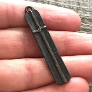 May include: A long, narrow, dark gray metal pendant with a weathered, matte finish. The object features a raised vertical ridge running down the center and a small circular loop at the top for attachment, displaying a simple, elongated rectangular shape.