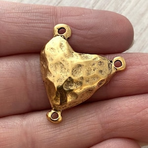 Könnte beinhalten: Ein goldfarbener herzförmiger Anhänger mit gehämmerten Texturen und drei kleinen Schlaufen zum Befestigen an Schmuck. Der Anhänger hat eine leicht unregelmäßige Form und ein warmes, antikes Finish.
