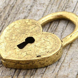 Può includere: Un lucchetto a forma di cuore, in stile antico e color oro. Il lucchetto presenta una serratura e un'ansa curva. La superficie presenta un disegno inciso, che gli conferisce un aspetto vintage. Il lucchetto è probabilmente in metallo.