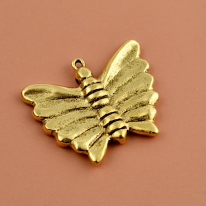 May include: A gold-colored butterfly pendant with detailed wing patterns and a loop for hanging. The butterfly has a textured, aged appearance, suggesting a vintage or antique style. The pendant is likely made of metal.