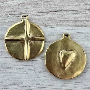 May include: Two antique gold-colored pendants. One pendant features a cross design, while the other has a heart shape. Both pendants have a loop at the top for hanging. The pendants have a rustic, textured appearance.