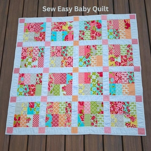 May include: A colourful baby quilt with a patchwork design featuring squares of various floral prints in shades of pink, red, blue, green, and yellow. The quilt is bordered with white and orange fabric.