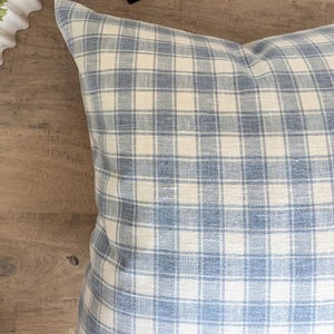 May include: A blue and white plaid pillow rests on a wooden surface. A black monkey statue and a white planter with green foliage are also visible. The pillow has a classic check pattern, adding a touch of traditional style to the decor.
