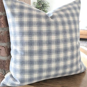 Pu&ograve; includere: Un cuscino quadrato con un motivo a quadri blu e bianco sporco. Il tessuto presenta una trama visibile con una griglia di linee orizzontali e verticali che si intersecano in un colore blu tenue su uno sfondo color crema chiaro.