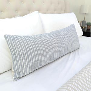 Peut inclure: Un long coussin lombaire rectangulaire en tissu texturé orné d'un motif de fines rayures verticales bleues et blanches. Le coussin est placé horizontalement sur un lit, appuyé contre deux grands oreillers blancs unis.