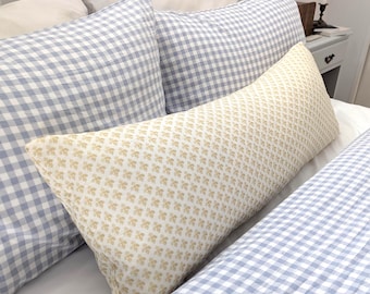 Può includere: Un lungo cuscino rettangolare in tessuto color crema con un piccolo motivo floreale beige ripetuto, posizionato davanti a due cuscini quadrati con motivo a quadretti vichy azzurro e bianco su un lenzuolo bianco.
