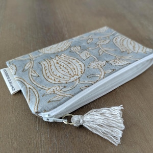 May include: A rectangular zippered pouch featuring a light blue fabric with a tan floral pattern. The pouch has a white zipper closure with a small beige tassel attached to the pull. A white fabric tag on the side reads COUSSINS ÉLÉGANCE.