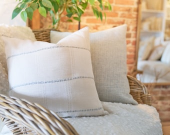 May include: Two square throw pillows resting on a woven wicker chair. The front pillow is off-white with a subtle grid pattern featuring thin, light gray horizontal and vertical lines. The back pillow is a solid, light gray textured fabric.