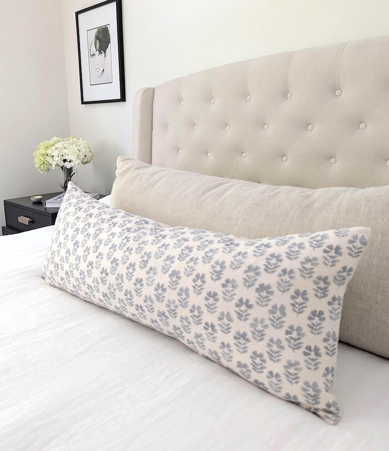 May include: A long, rectangular decorative pillow with a floral pattern in shades of gray and blue rests on a white bed. The pillow is in front of a beige bolster pillow and a tufted headboard. A framed picture hangs on the wall.