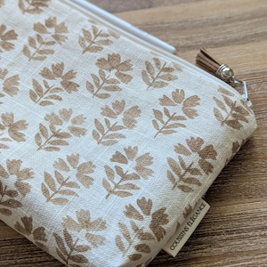 May include: A cream-colored pencil case with a repeating pattern of brown floral designs. The case has a zipper closure with a brown tassel and a silver-colored metal pull. The label reads "COUSSINS ÉLÉGANCE".