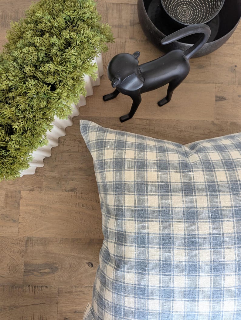 May include: A blue and white plaid pillow rests on a wooden surface. A black monkey statue and a white planter with green foliage are also visible. The pillow has a classic check pattern, adding a touch of traditional style to the decor.
