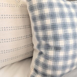Pu&ograve; includere: Due cuscini quadrati in tessuto testurizzato. Il cuscino anteriore presenta un motivo a quadri blu e bianco sporco, mentre quello posteriore &egrave; in tessuto color crema con righe orizzontali di piccole linee blu tratteggiate. &Egrave; visibile un'etichetta.