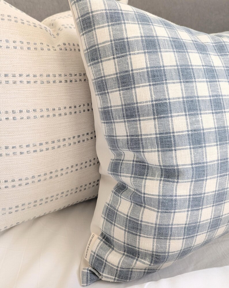 Pu&ograve; includere: Due cuscini quadrati in tessuto testurizzato. Il cuscino anteriore presenta un motivo a quadri blu e bianco sporco, mentre quello posteriore &egrave; in tessuto color crema con righe orizzontali di piccole linee blu tratteggiate. &Egrave; visibile un'etichetta.