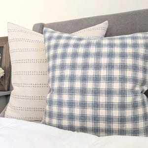 Pu&ograve; includere: Due cuscini quadrati posizionati su un letto. Uno presenta un motivo a quadri blu e bianchi, mentre l'altro &egrave; in tessuto color crema con righe orizzontali di piccole linee tratteggiate marroni. Entrambi i cuscini hanno un aspetto morbido.