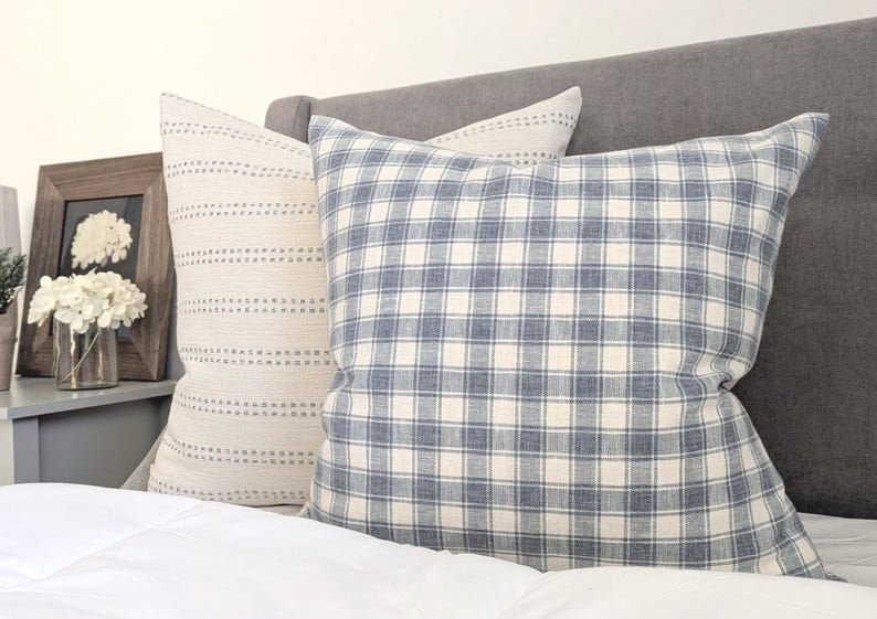 Pu&ograve; includere: Due cuscini quadrati posizionati su un letto. Uno presenta un motivo a quadri blu e bianchi, mentre l'altro &egrave; in tessuto color crema con righe orizzontali di piccole linee tratteggiate marroni. Entrambi i cuscini hanno un aspetto morbido.