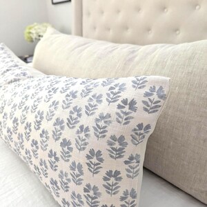 May include: A long, rectangular pillow with a light beige background and a repeating pattern of blue floral sprigs. The pillow is placed on a white surface, with a beige headboard in the background.