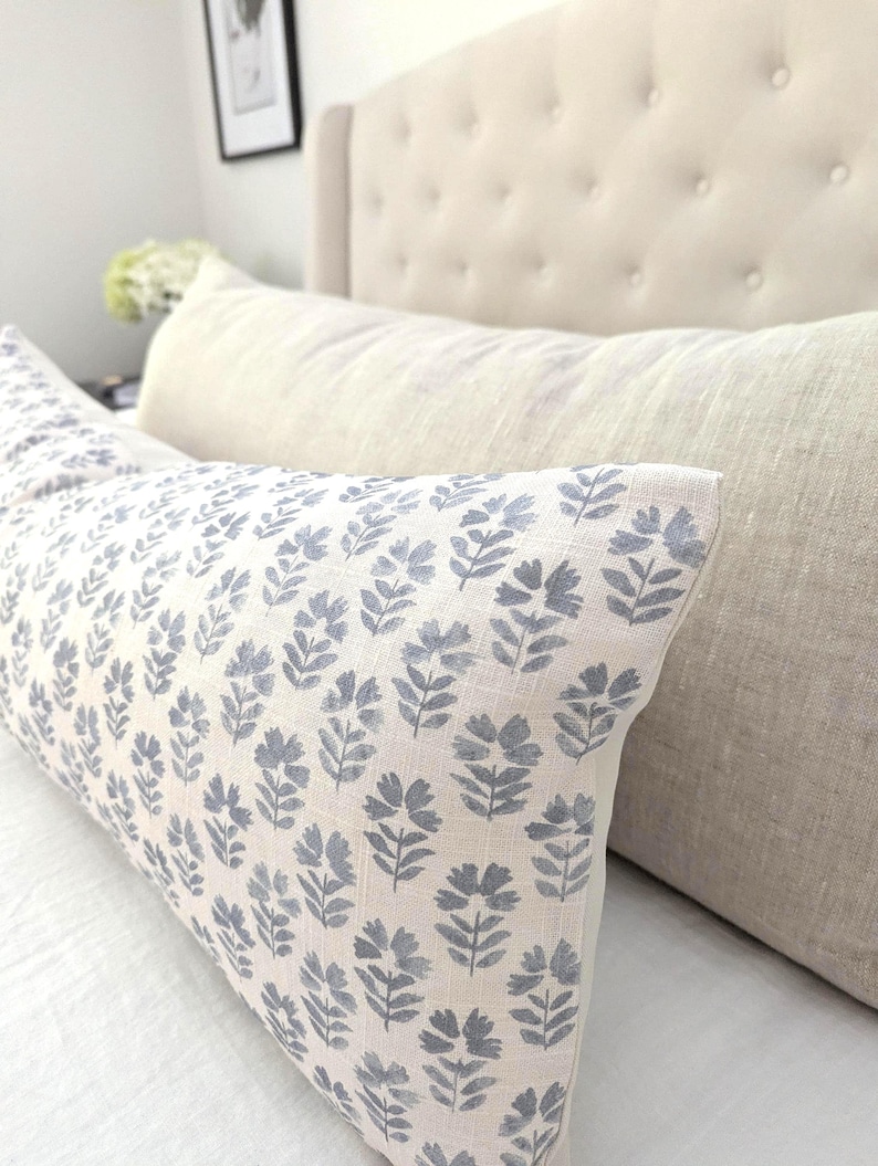 May include: A long, rectangular pillow with a light beige background and a repeating pattern of blue floral sprigs. The pillow is placed on a white surface, with a beige headboard in the background.