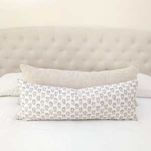 May include: A decorative pillow with a blue floral pattern on a white background. The pillow is rectangular and rests on a white bed with a beige headboard. A second, solid-colored pillow is behind it.