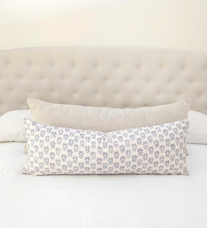 May include: A decorative pillow with a blue floral pattern on a white background. The pillow is rectangular and rests on a white bed with a beige headboard. A second, solid-colored pillow is behind it.