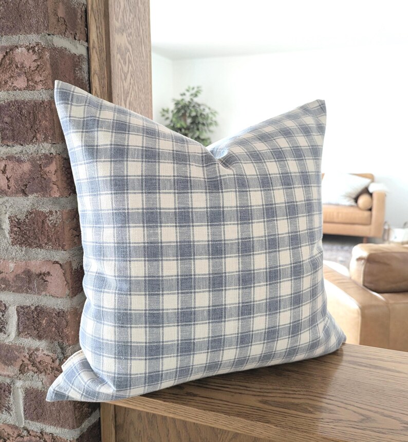 Pu&ograve; includere: Un cuscino quadrato con un motivo a quadri blu e bianco sporco. Il tessuto presenta una trama visibile con una griglia di linee orizzontali e verticali che si intersecano in un colore blu tenue su uno sfondo color crema chiaro.