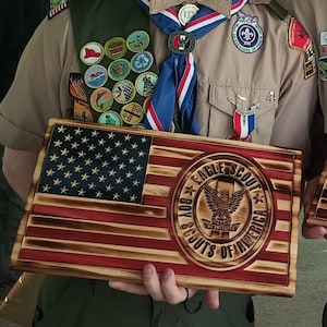 May include: A wooden American flag with red and white stripes and blue stars. The flag has a circular emblem with an eagle and the text "EAGLE SCOUT BOY SCOUTS OF AMERICA". The flag is held by a person wearing a Boy Scout uniform.