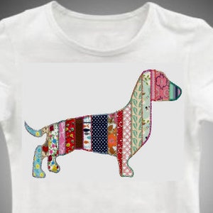 May include: White t-shirt with a dachshund dog design. The dog is made up of colourful fabric strips in red, blue, pink, and green. The t-shirt has a crew neck and short sleeves. The dog design is a patchwork style.