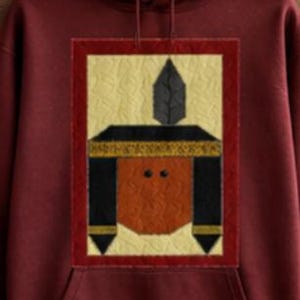 May include: A maroon hooded sweatshirt featuring a rectangular graphic print on the chest. The graphic depicts a stylized figure with a brown face, black hair, a gold headband, and a single black feather, all set against a cream background.