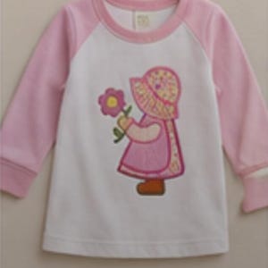 Peut inclure: Un t-shirt à manches longues avec un torse blanc et des manches raglan rose clair. L'avant présente une broderie représentant une silhouette portant un bonnet à motifs et une robe rose, tenant une fleur violette au centre jaune et des chaussures marron.