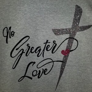 以下が含まれることがあります： グレーのTシャツに、黒いグリッターの十字架と「No Greater Love」という文字、そして小さな赤いハートが描かれています。
