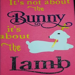以下が含まれることがあります： ピンクのTシャツに、キラキラの文字で「It's not about the Bunny it's about the Lamb」と書かれています。Tシャツには、金色の角を持つ鹿の白い紙の切り抜きが貼られています。