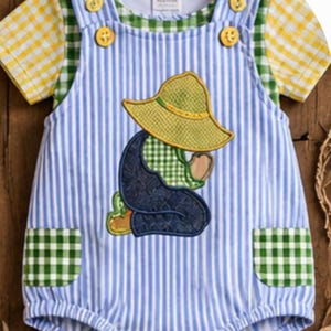 May include: A blue and white striped romper featuring yellow and green gingham sleeves and pockets. The front displays an embroidered applique of a figure in a yellow hat and dark blue overalls. The garment has yellow buttons at the shoulders.